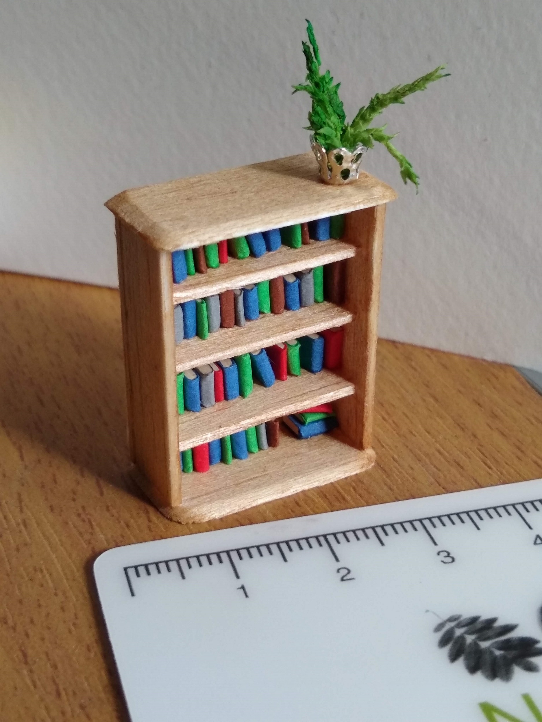 Quarter scale bookcase, made in light coloured wood, brightly coloured books on the shelves and a small plant in a silver filigree pot on top