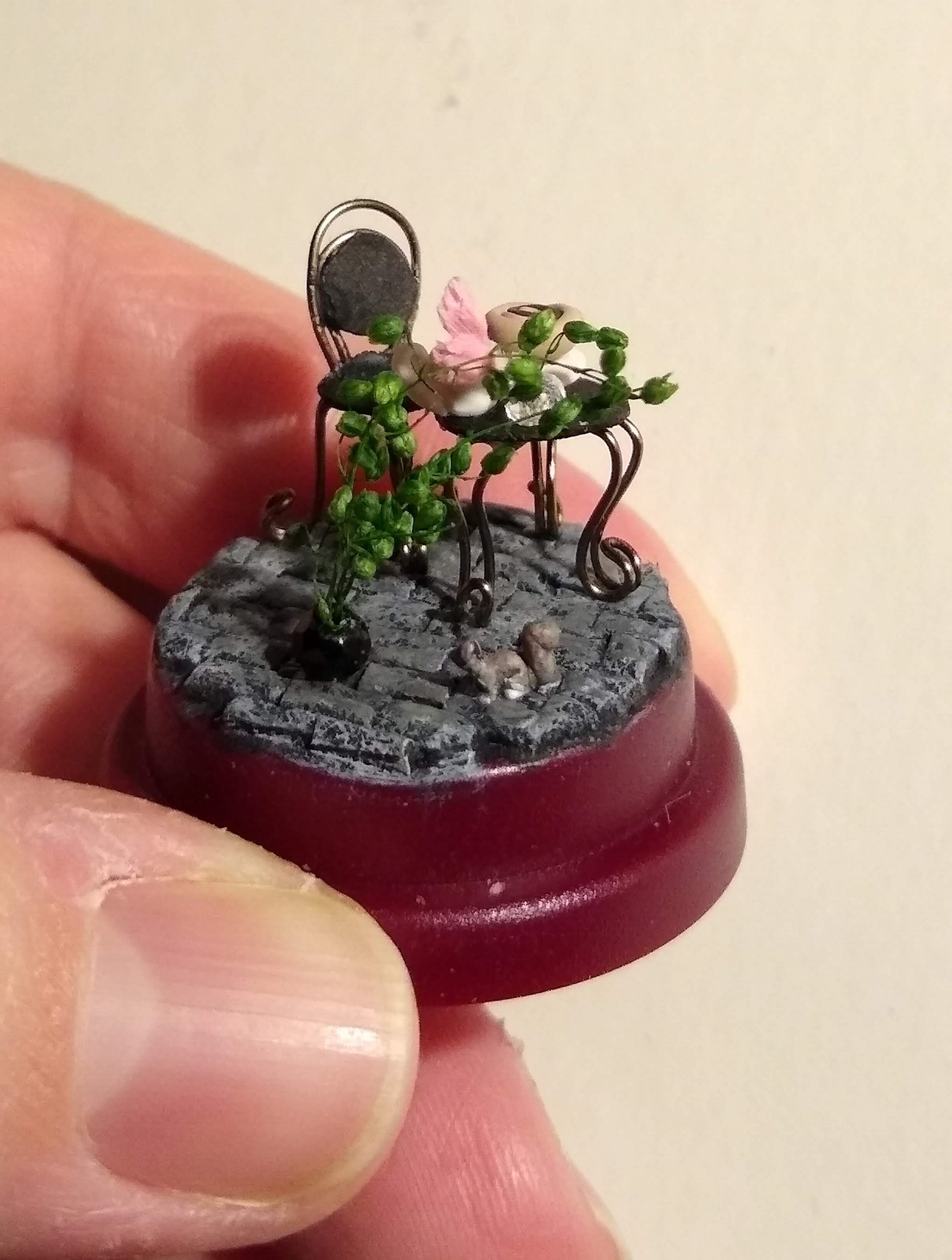 A tiny cafe scene in a thimble case. It is a table and chair with a plant in a pot nearby. There is a tiny squirrel on the ground and a cinnamon roll on a plate on the table.