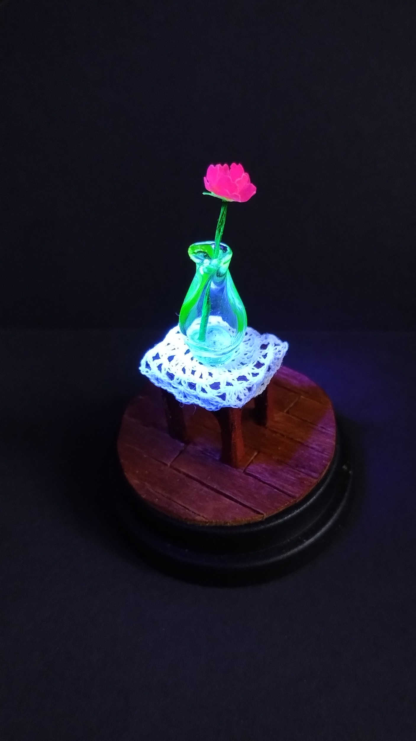 A small uranium glass vase glows brightly along with the white crochet doily underneath and pink flower in the vase, on top of a small wooden side table.