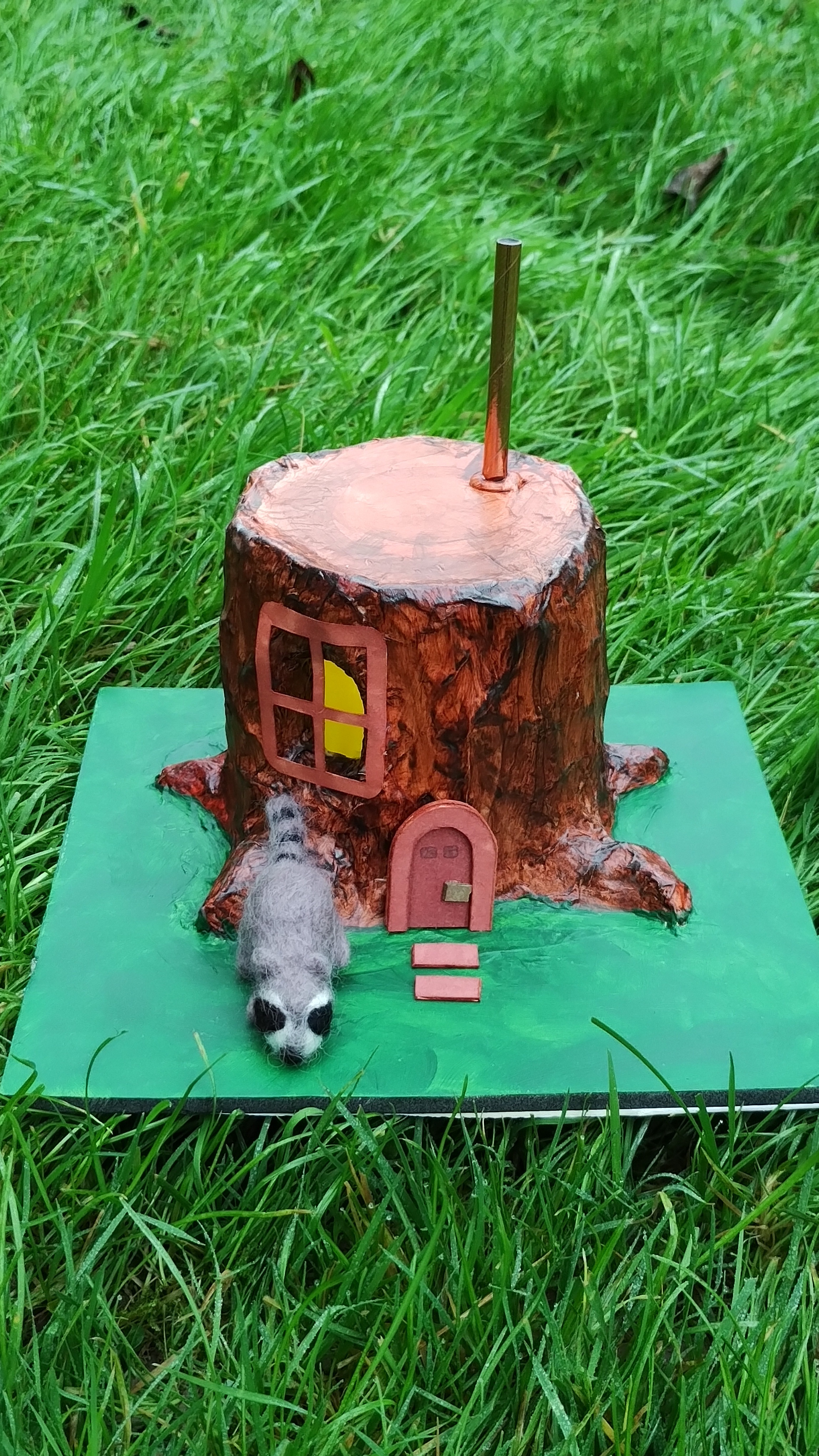 diorama with a needlefelted racoon outside of a house made from a tree stump