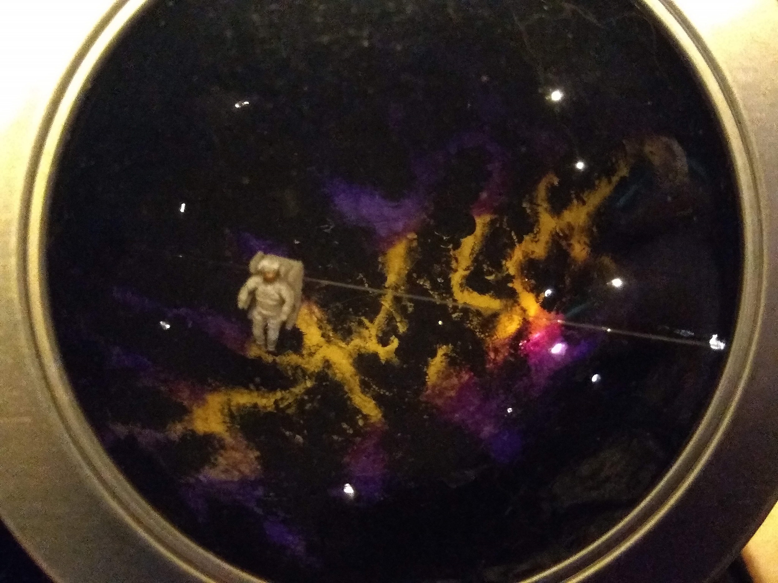 A 1:144 scale model man in a spacesuit floats above a brightly lit whispy purple and yellow galaxy and lots of stars.