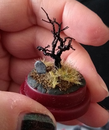 a hand holding a very tiny diorama in a thimble case, of a spooky looking tree, some dead grass and a tiny tombstone