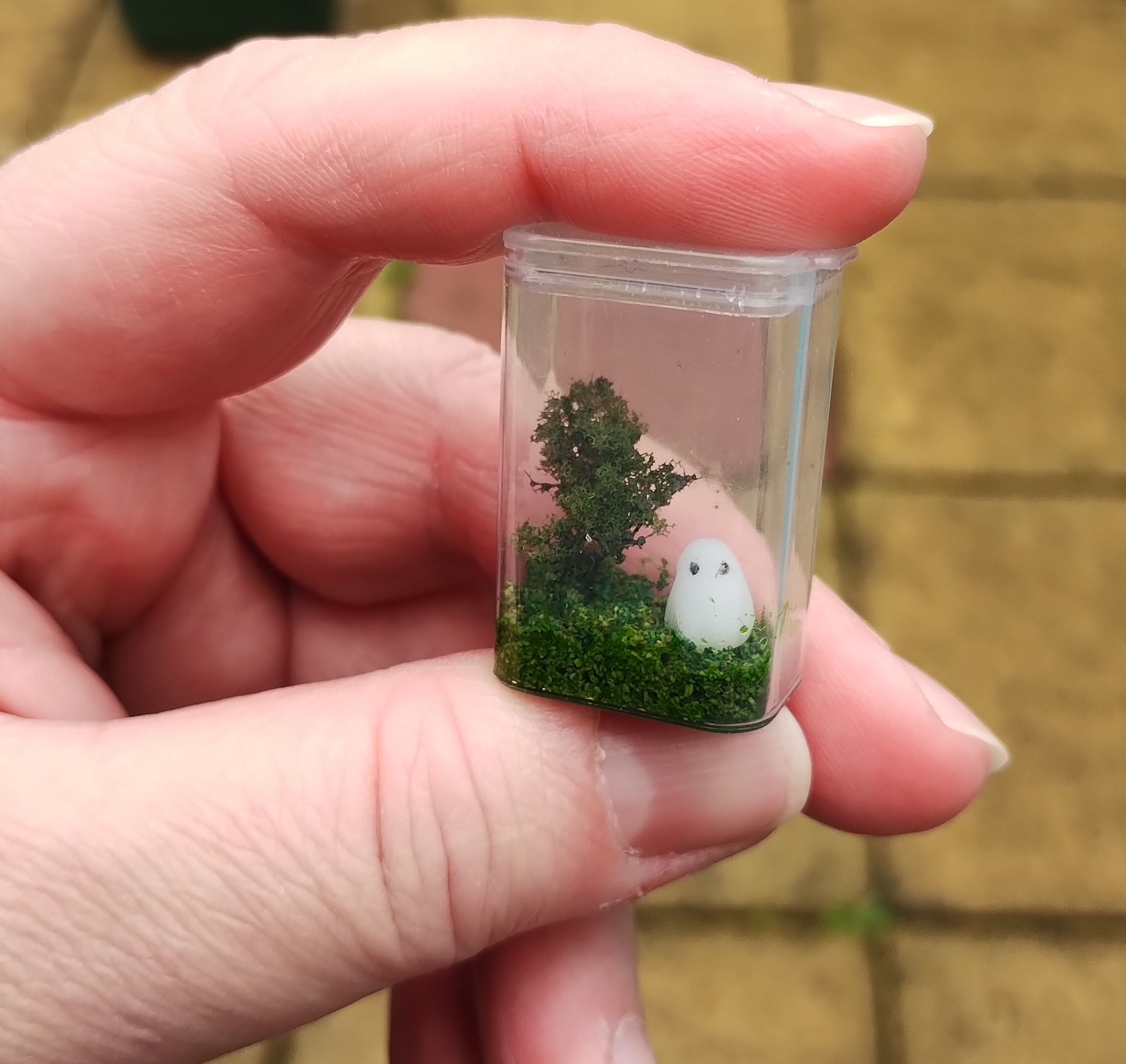 A hand holding up a miniature tictac box with a diorama of a ghost by a tree on some greenery