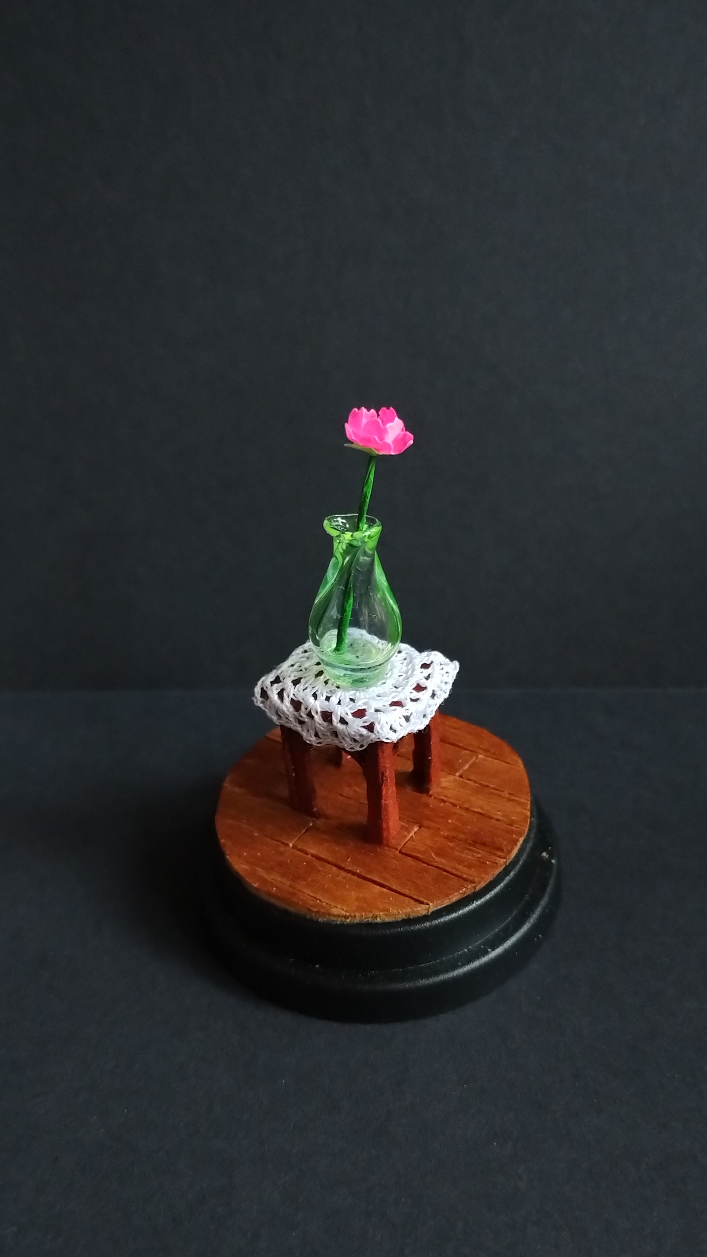 The flower in a green glass vase on a white doily, on a small side table in normal light.