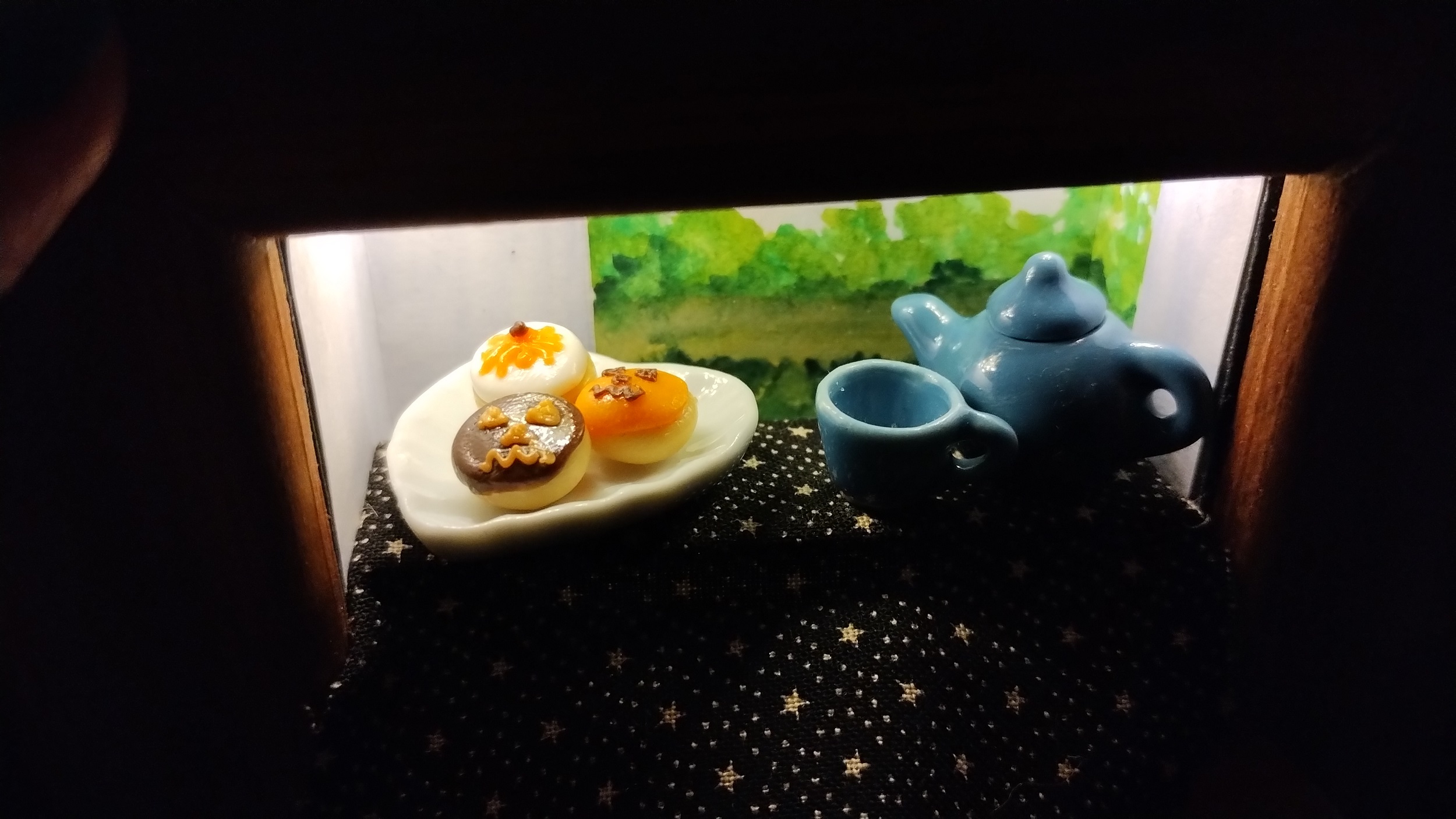 Detailed close up photo of the tiny diorama of a table by an open window, brightly lit. The table has a blue teapot and cup on it along with a plate of doughtnuts decorated for halloween with orange icing and jack o lantern face