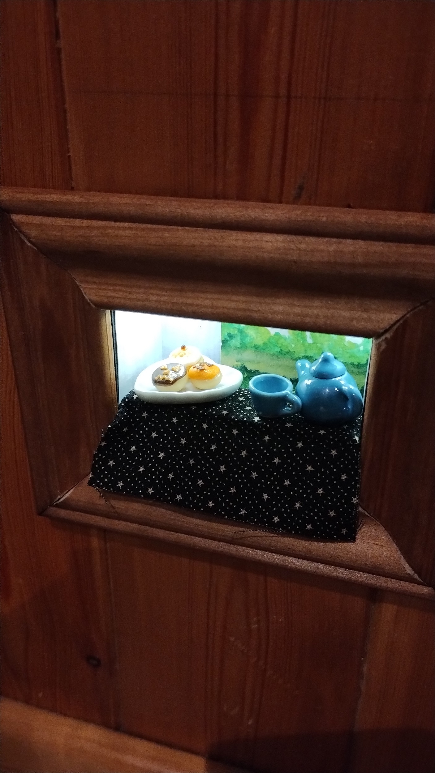 a tiny diorama of a table by an open window, brightly lit. It is framed with a golden brown wood trim and set into a wood panelled wall.